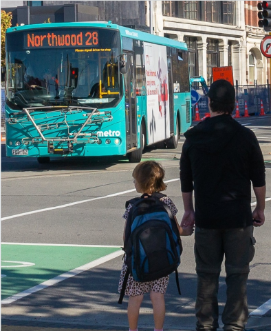 Father and daughter wait to cross the road, with a bus in the background